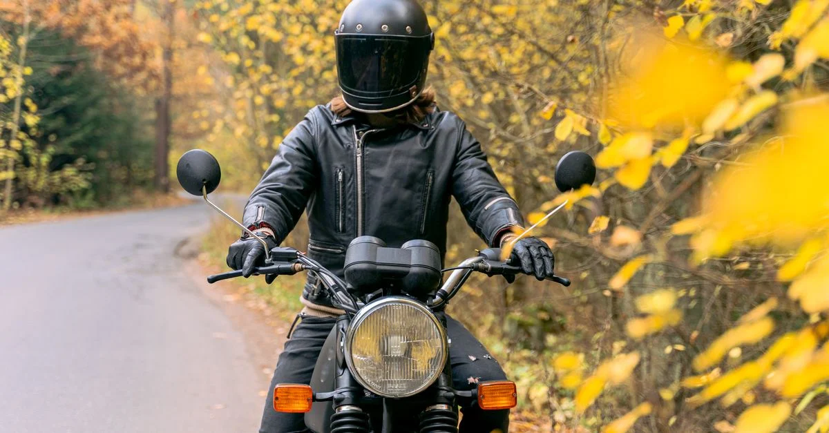 Un motard sur une moto, vêtu d'une veste noire et d'un casque, roule sur une route forestière pittoresque en automne.